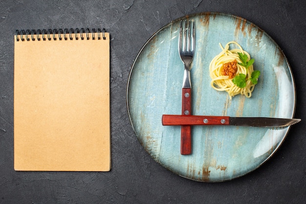 Above view of cutlery set pasta meal with meat on blue plate and notebook on black background with free space