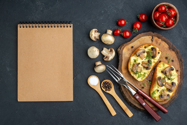 Above view of delicious snack with mushrooms cutlery set on wooden board and spices raw vegetables notebooks on black background