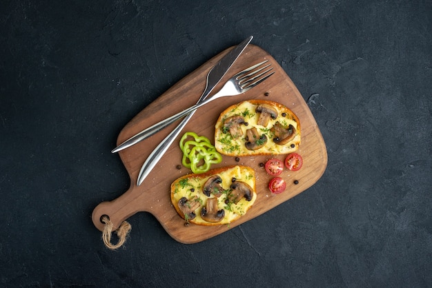 Above view of delicious snacks with mushroom fresh vegetables and cutlery set on wooden cutting board on black background