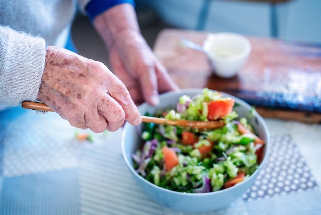 View of elderly woman's hands stirring the salad with a wooden spoon on the table of the kitchen