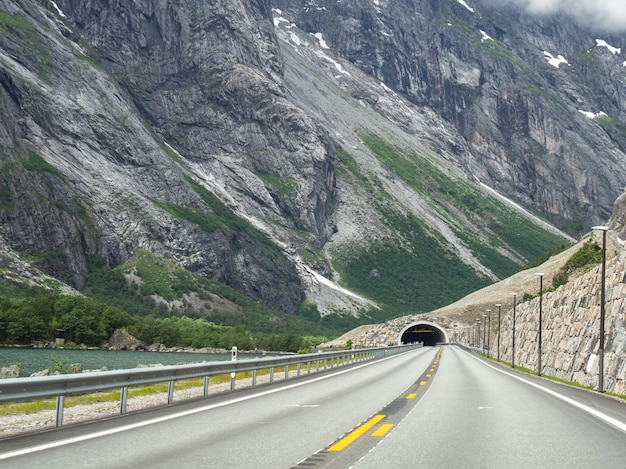 View of an empty asphalt road with double yellow lines and a tunnel in the mountain by the lake. Norwegian mountain road in cloudy weather