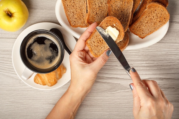 View of female hands spreading butter on toast with cup of black coffee