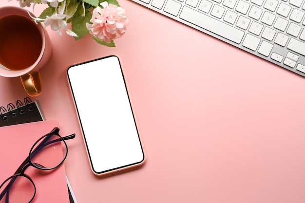 Above view of female workspace with mobile phone keyboard glasses coffee cup and notebook on pink pastel background