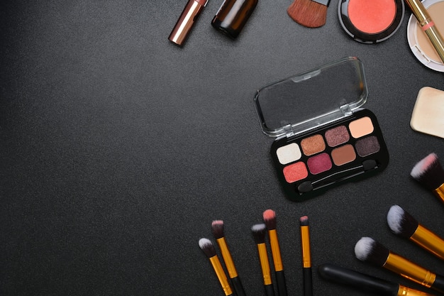 Above view of feminine desk with with lipsticks, eye palette, face powder, brushes and copy space on dark background.