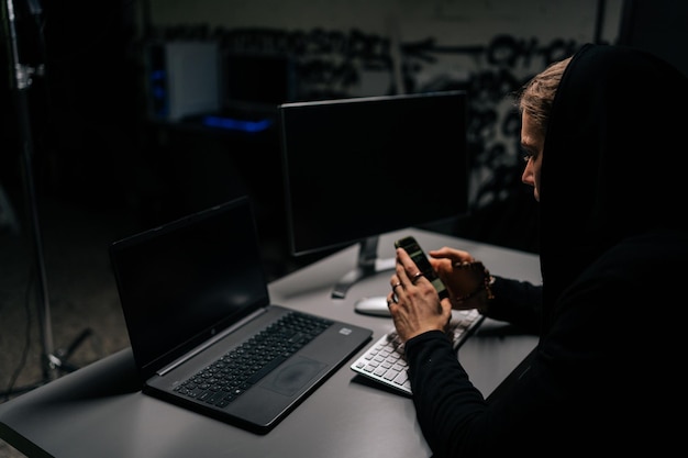 View from back to hidden hacker man wearing sweatshirt with hood engaged in hacking into security systems using mobile phone