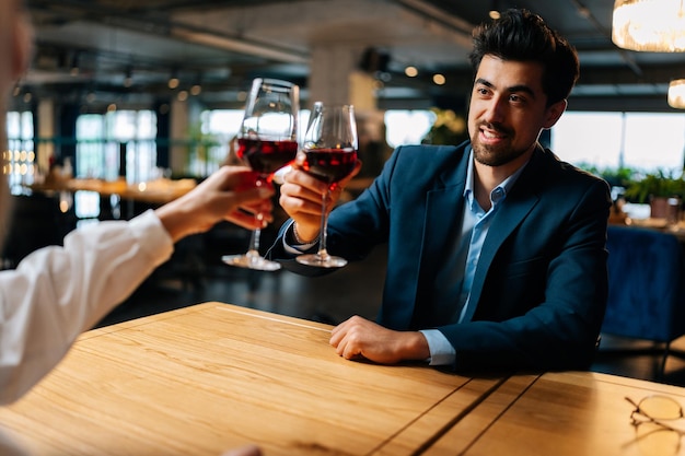 View from back of unrecognizable female clinking glasses of red wine with smiling elegant man in suit sitting at table in fancy restaurant at evening