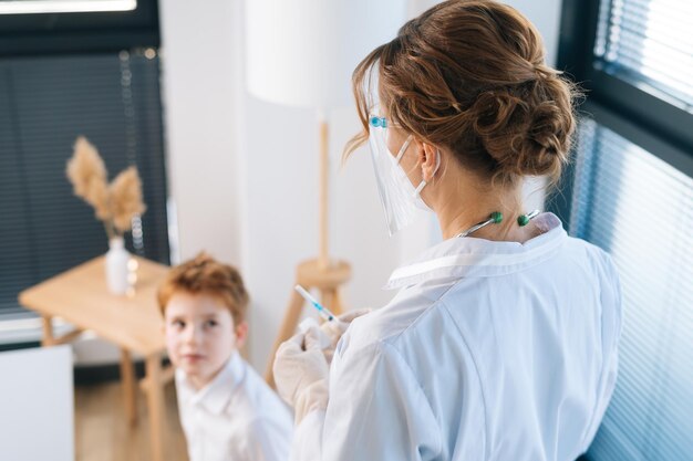 View from back of unrecognizable female doctor in latex gloves preparing vaccine for child boy at hospital with light interior