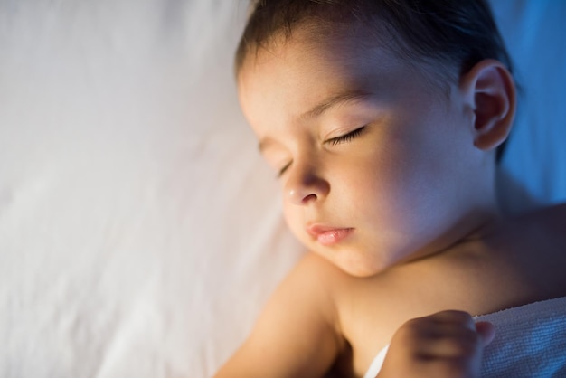 View from above closeup portrait of adorable baby girl sleeping in white bed at night Little toddler taking a nap in dark room in lamplight Bed time for kids Bedroom and nursery interior