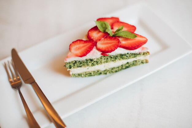 View from above of delicious cake with cream and strawberries in cafe. Tasty creamy dessert serving with knife and folk on white plate on table. Concept of confectionery and pastry.