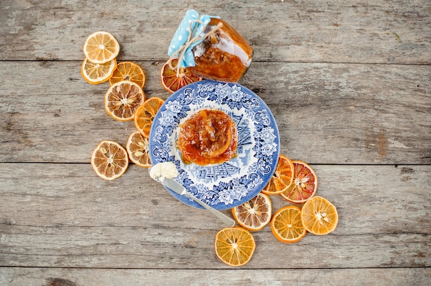 View from above on the delicious orange jam on bread served on the blue ornate plate near the glass jar and dried oranges on the wooden table