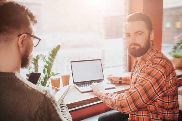 A view from a different angle where on of hipsters is sitting and looking to the man with computer while that man is looking straight ahead and putting his right arm over the keyboard.