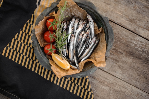 View from above on the fresh anchovies on the plate with a rosemary, cherry tomatoes and lemon on the wooden table and black and yellow napkin