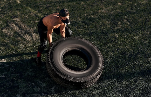 Photo view from above of handsome fitness muscular male in mask to increase load on breathing muscles doing exercises on big tire outdoors copy space for text shirtless sportsman doing workout on stadium