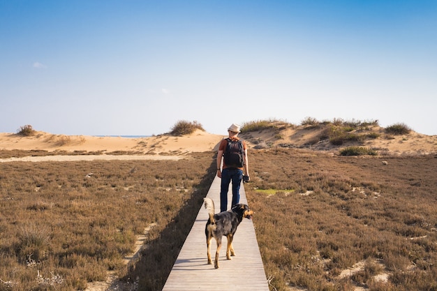 View from behind of a man walking with his dog on a road leading through beautiful landscape.