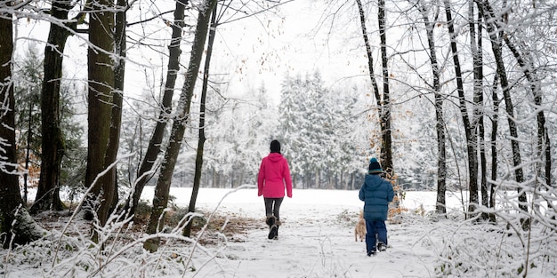 View from behind of a mother and her son walking in beautiful snowy forest with their cute small dog around them.