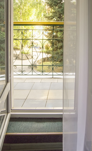 View from the room to the terrace or balcony with a metal railing. The sun shines through a transparent curtain and trees during sunset, vertical shot