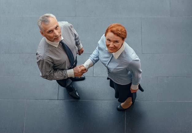 Photo view from above of smiling woman and mature man in business suits standing next to each other shaking hands and looking at camera