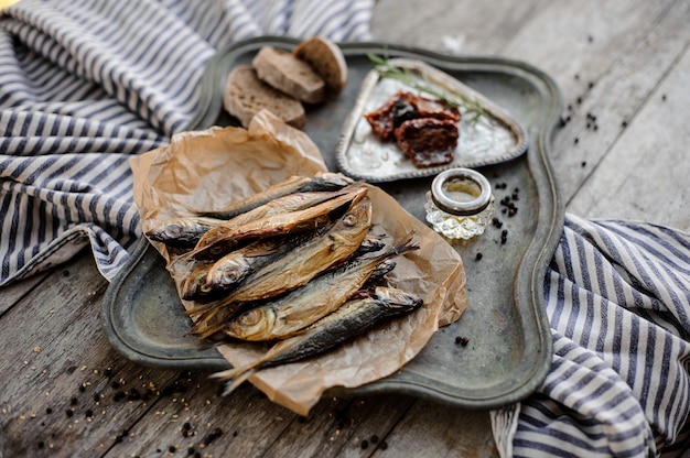 View from above on the tasty smoke-dried horse mackerel fish on the paper on the metal tray with a sun dried tomatoes, oil and bread on the grey striped napkin on the wooden table.