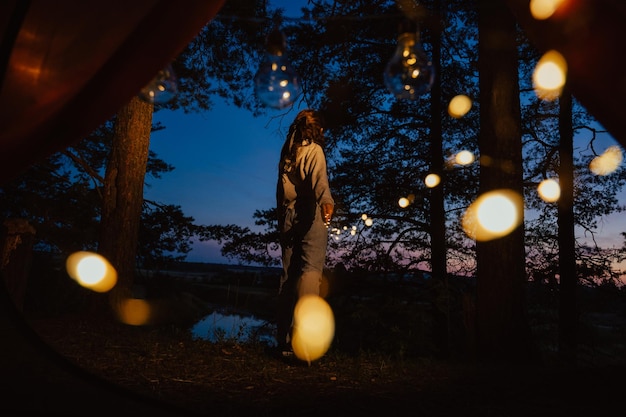 View from the tent A young woman stands tall and holds lights in her hands against the background of trees and water Outdoor recreation The magic is in the picture