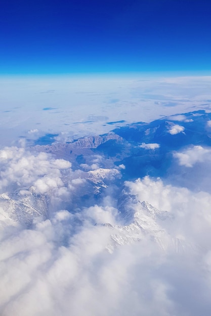 View from window of plane to blue sky and snowy mountain peaks in white clouds