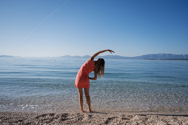 View from behind of a young woman standing ankle deep in calm morning sea water stretching with her arm to the side.