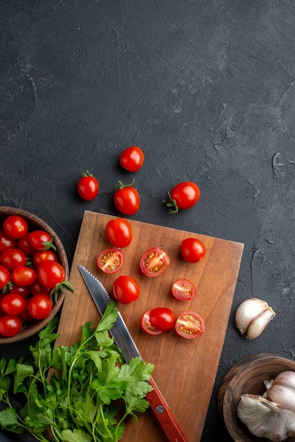 Above view of green bundle fresh tomatoes on wooden cutting board and in bowl on black distressed surface