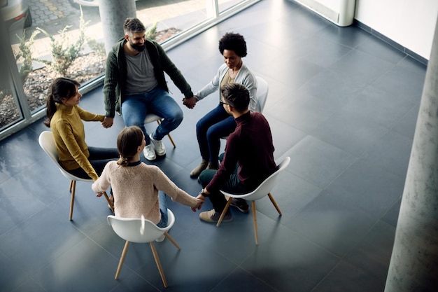 Above view of group therapy attenders holding hands while sitting in a circle