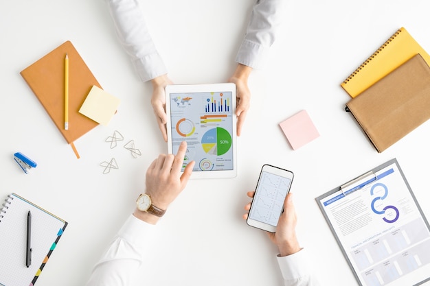 View of hands of two analysts or brokers with gadgets discussing financial data on touchscreen while one of them pointing at circle diagram