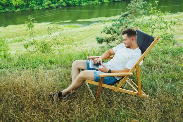 View of handsome guy lying on chair in nature and working with laptop resting alone thoughtfully looking up A man on a trip in the countryside the concept of people's lifestyle