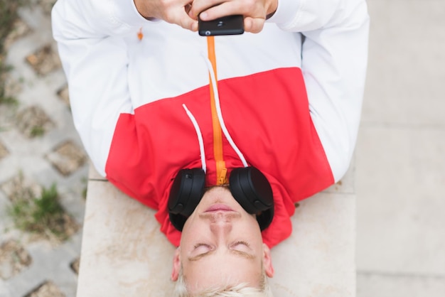 Above view of handsome young blond Caucasian man lying on the floor outside with bluetooth headphones on the neck listening the music and texting messages Lifestyle people and music concept