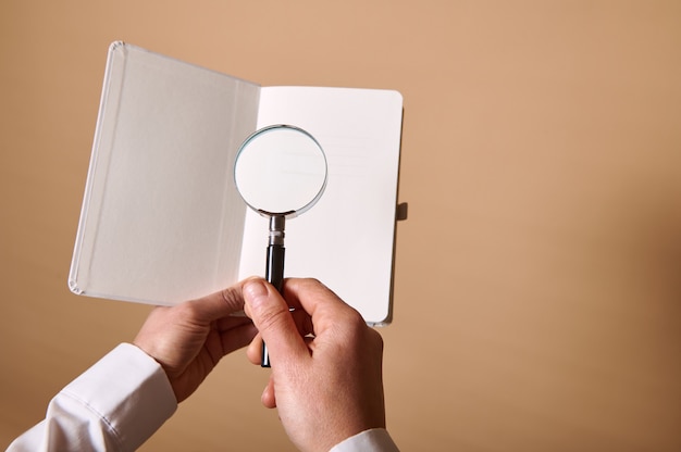 A view of an image of white blank sheet with a magnifier or magnifying glass in female hands on beige wall with copy space