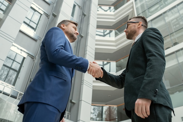 Below view of male lawyers in formal wear standing in hall of commercial center and making handshake as symbol of partnership