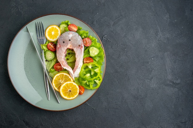 Above view of raw fish and fresh vegetables lemon slices and cutlery set on a gray plate on the left side on black surface with free space