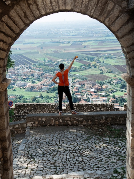 A view through the arch of a girl who stands with his back in the center of the photo and looks from above at the roofs of houses and fields, his hand is raised up, an orange is in his hand