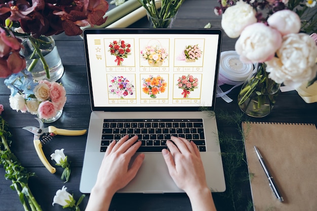 Photo above view of unrecognizable florist standing at desk with beautiful flowers and hand tools and typing on laptop keyboard while making online order of flowers for own shop