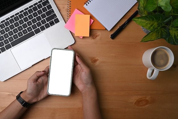 Above view woman holding mock up mobile phone with blank screen on her office desk.