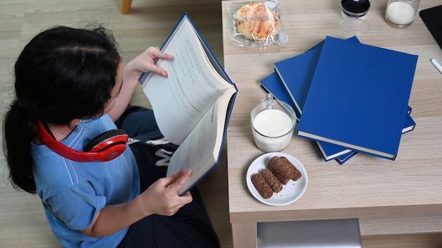 Photo above view of young asian girl reading book while sitting on floor in living room