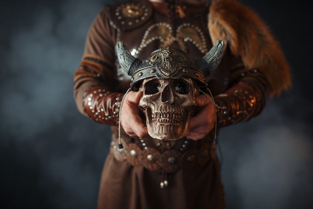 Viking dressed in traditional nordic clothes holds human skull in helmet