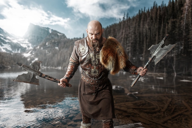 Viking with axes in hands dressed in traditional nordic clothes standing at the lake. Ancient warrior at the river, rocky mountains