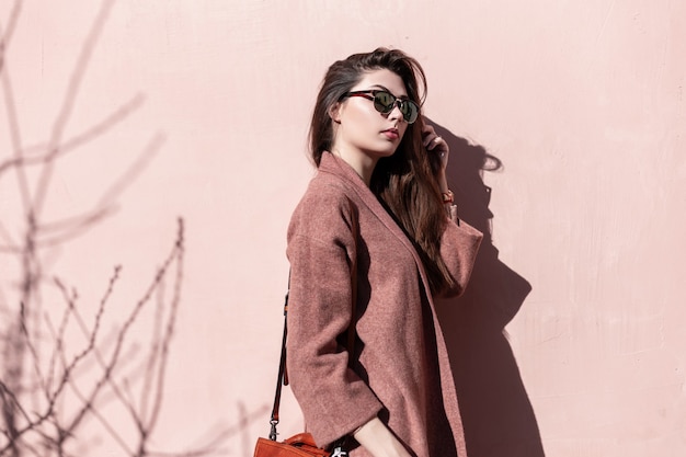 Vintage portrait attractive lovely young woman in dark stylish sunglasses with long hair in elegant coat with handbag near vintage pink wall outdoors. Beautiful gorgeous girl model enjoys spring sun.