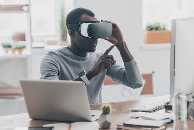 Virtual work. Handsome young African man in VR headset pointing in the air while sitting at the desk in creative office