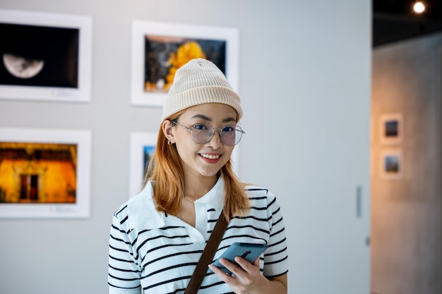 Visitor woman smiling at picture art gallery collection in front framed paintings pictures she holding mobile phone