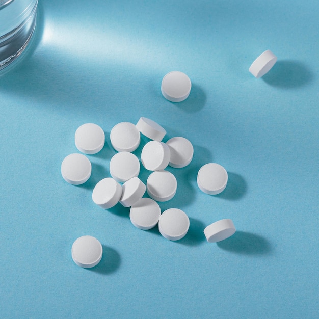 Vitamin white pills freely laid near glass of water on light blue background