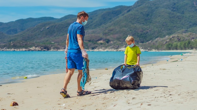 Volunteer blue face mask lazur sea sand beach Son helps father hold black bag for pick up garbage Problem spilled rubbish trash planet pollution environmental protection Natural child education