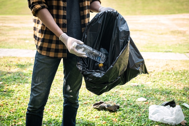 Volunteer man in gloves to picking up plastic bottle into plastic black bag for cleaning the park during environmental activity to collecting garbage