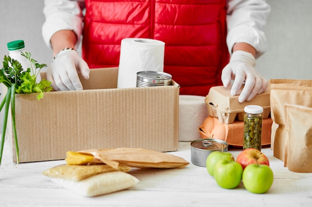 Volunteer in the protective medical mask and gloves putting food In donation box. Delivery man employee in red vest packing box with food. Service quarantine pandemic coronavirus.