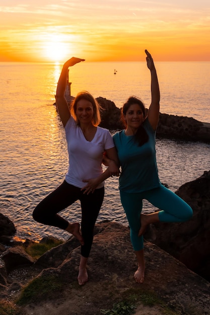 Vrksasana two women doing meditation and yoga on a rock at sunset next to a lighthouse in the sea healthy and naturist life