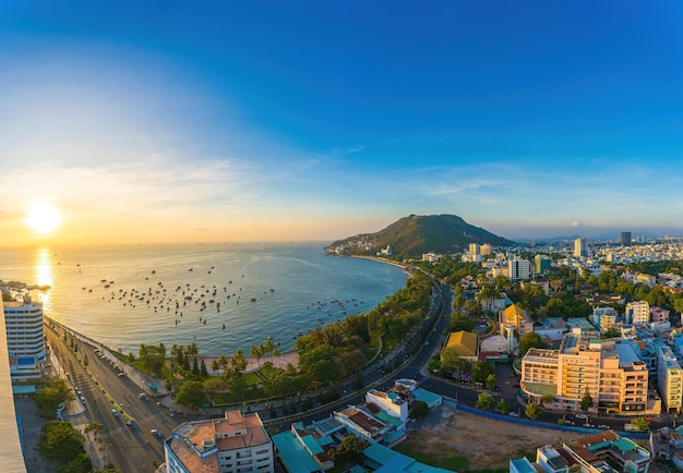 Vung Tau city aerial view with beautiful sunset and so many boats Panoramic coastal Vung Tau view from above with waves coastline streets coconut trees and Tao Phung mountain in Vietnam