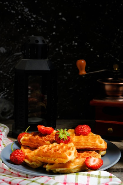 Waffles with strawberries and honey sweet pastries on plate against background coffee grinder and decorative lamp with candle