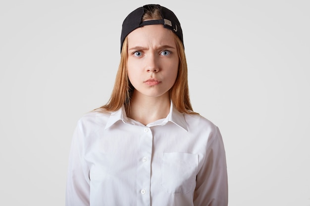 Waist up grumpy female model with sullen expression, wears black cap and elegant shirt, being dissatisfied with something, isolated on white. People and emotions